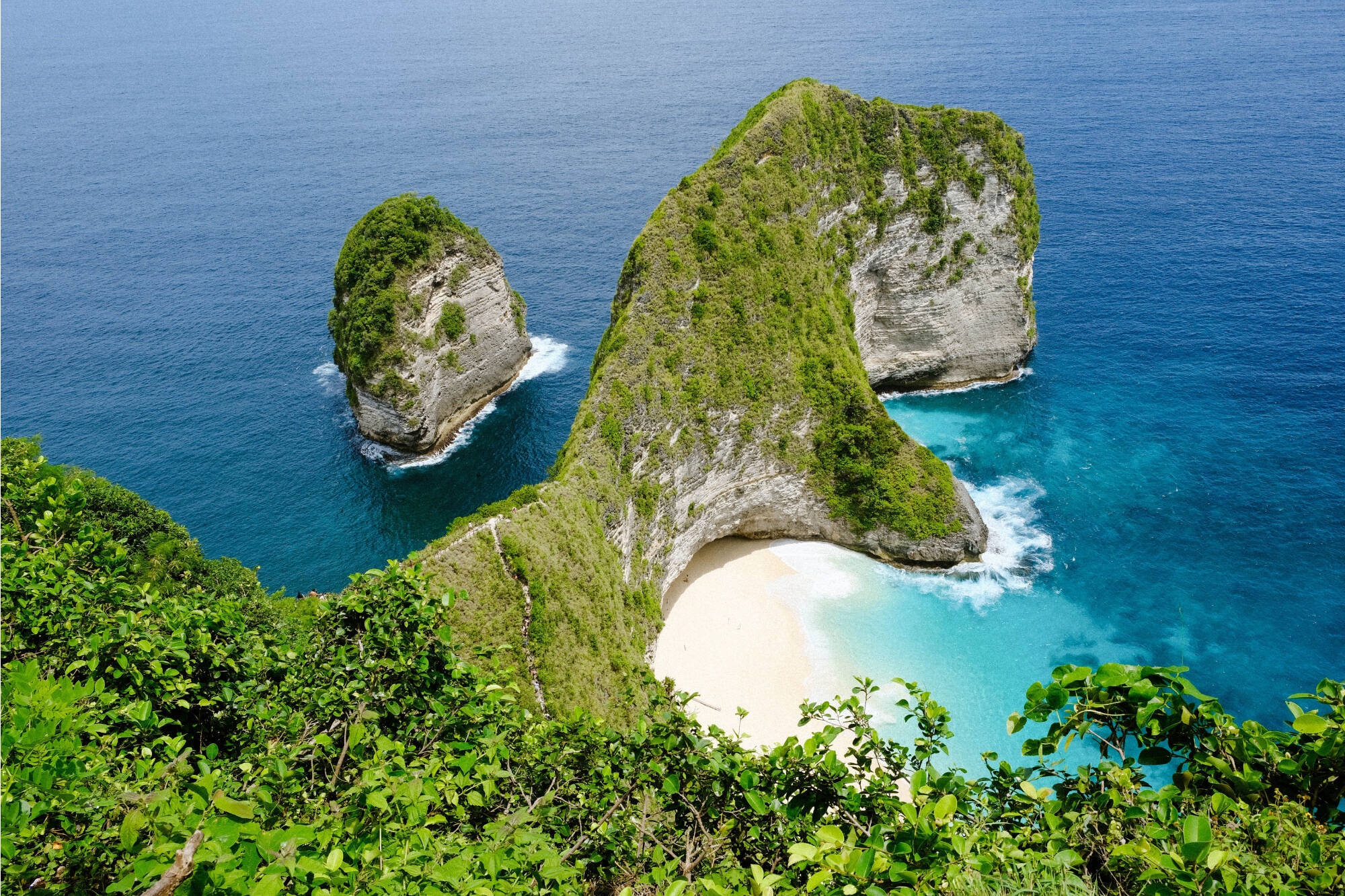 A stunning view of Kelingking Beach on Nusa Penida Island, Indonesia, featuring dramatic limestone cliffs covered in greenery, turquoise waters, and a secluded white sand beach framed by the Indian Ocean.